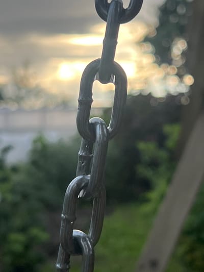 The image shows a close-up of a metal chain suspended against a blurred background. The chain links are dark and appear to be made of iron or steel. The background shows a hazy, bright sky with what looks like the sun shining through clouds or trees. There are also blurry green shapes that suggest foliage. A light-colored, vertical pole is partially visible on the right side of the frame. The overall impression is of an outdoor scene, possibly in a park or a yard. The location is specified as Бучацінскі сельскі Савет, Belarus.