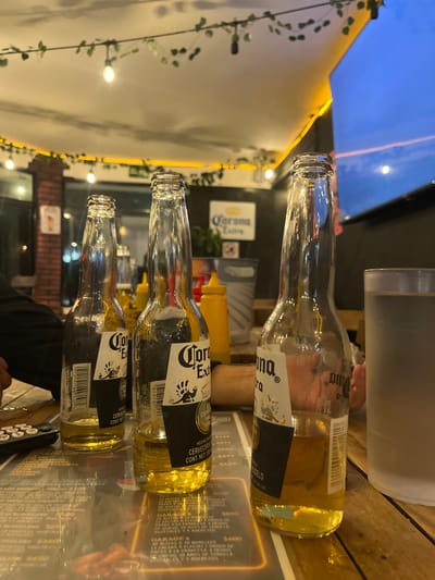 Three Corona Extra beer bottles, partially consumed, lie on a reflective surface. The topmost bottle has the least amount of beer remaining. A cooler and condiment bottles are in the background. String lights adorn the outdoor space, visible on the left side. A television screen displays the sky above. The reflective surface shows a menu underneath.