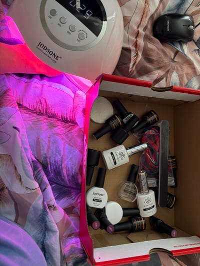 The image captures a casual, indoor setting in Bearpark, United Kingdom, featuring a collection of nail care essentials and a computer mouse on a patterned fabric.
A white JODSONE SUNX20 Max Nail Lamp occupies the lower left, emitting a distinct pink-purple light. Its digital display shows "29" (likely seconds remaining), and it has buttons for "10S", "30S", "60S", and "99S" (Low Heat Mode). An open cardboard box, marked with "52-22N5W8 265x210x100" on its side, is filled with various gel nail polish bottles and tools. Brands visible include "BORN PRETTY" (with "BASE GEL" and "TOP COAT"), "Shining She" ("SOAK-OFF UV&LED Gel Polish"), "JODSONE" ("Gel Nail Polish Top Coat" and "Nail Dehydrator"), and "JIAZHIGAI". Some bottles are labeled with color codes like "A382", "A383", and "A384". A black nail file, red string or band, and white cotton pads are also scattered within the box.
In the upper left, a black wired Microsoft computer mouse rests on the fabric. The entire scene is laid out on a soft, patterned fabric, possibly bedding or a blanket, adorned with abstract floral or leaf designs in shades of pink, purple, brown, and cream. The presence of the nail lamp and numerous polishes suggests a home manicure or nail art session is either underway or has recently concluded.