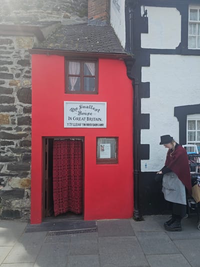 A medium shot reveals "The Smallest House in Great Britain" nestled in the city of Llandudno Junction, United Kingdom. The tiny structure is painted a vibrant red. A woman with a black hat is standing outside of the building. Next to the house on the left is a stone wall, and on top is a shingled roof. Directly in front of the house is an entry space with a brown frame and a red checked fabric inside. A window is to the left of the entryway, and another, smaller window is to the right. A sign to the left of the door reads, "The Smallest House In Great Britain. Y TY LLEIAF YM MHRY DAIN FAWR." The building next to the house is white and black. The street is to the right of the house.