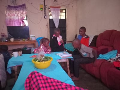 This image depicts an indoor scene, likely a living room, in Kiminini, Kenya. Five children are present in the room.

The environment includes light-colored walls, a wooden table covered with a bright blue tablecloth in the foreground, and a red-orange upholstered sofa on the right. A flat-screen television rests on a wooden stand against the left wall, beneath a window with purple and white patterned curtains. A doorway or window with a white sheer curtain and security bars is visible at the rear center. Electrical wiring runs along the wall, connecting to a yellow power meter with a green indicator light. The floor is dark.

On the blue tablecloth, a yellow bowl filled with green leafy vegetables and other food items is centrally placed, with a clear plastic bag containing additional items nearby. A white mug is on the table, and another is on the television stand. A red and black plaid blanket is draped in the lower foreground.

Of the five children, one is partially visible on the far left, seated near the table. A young girl in a sequined top sits on a blue plastic chair at the middle left, looking towards the table. Another young child in a black and pink top stands or kneels at the table, manipulating an object. On the sofa, a young boy in a red and blue top is seated, looking forward, while another young boy in a light gray top and dark pants is seated with bent legs, looking down towards the table area. The children are positioned around the table and sofa, appearing engaged with their immediate surroundings.