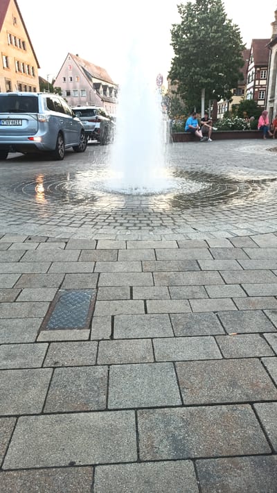 In Altdorf bei Nürnberg, Germany, a paved public square features a prominent water fountain in the center, with water spraying upwards. The square is made of grey cobblestones with distinct circular patterns formed by the splashing water. On the left, a light blue SUV with a German license plate is parked. In the background, traditional German architecture with cream-colored facades and red-tiled roofs lines the square. Several individuals are seated on a low wall or bench to the right of the fountain, with some appearing to be conversing or looking at their phones. A large green tree with dense foliage stands behind the seated individuals. The lighting suggests it is late afternoon or early evening.