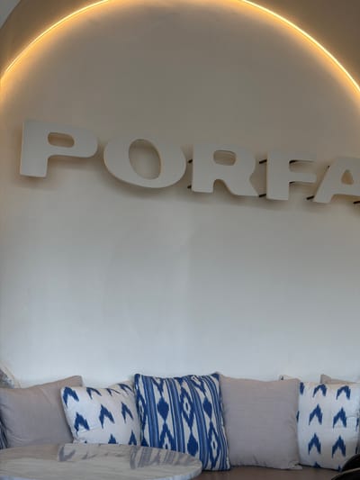An interior shot shows a wall with large white cutout letters spelling "PORFA". The letters are mounted on the wall with black pegs. A curved light fixture is mounted above and behind the letters, casting a warm glow on the wall. In the foreground, a sofa with several decorative pillows is visible. The pillows are upholstered in light-colored fabric, with two featuring a blue and white geometric pattern. A portion of a round table with a marble-like surface is in the lower left corner. This scene is located in Oakland Park, United States of America.