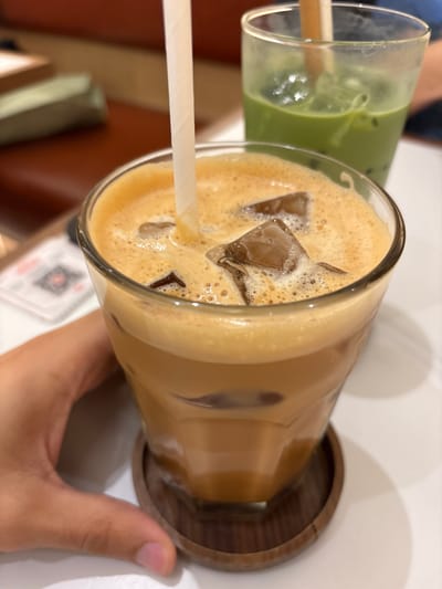 A hand, with tanned skin and short fingernails, grips the base of a glass tumbler containing an iced beverage. The beverage is a light brown, frothy liquid with visible ice cubes and a white straw. Behind this glass, another tumbler is visible, filled with a green liquid, also containing ice. The tumblers sit on a white surface, with a dark wooden coaster under the foreground glass. In the background, a blurred reddish-brown chair is visible, with a green and khaki object resting on it. A white card with a QR code is partially visible on the left. The scene appears to be set indoors, possibly in a cafe, and judging by the style and presentation, it could be in Ipoh, Malaysia, a city known for its vibrant cafe culture.