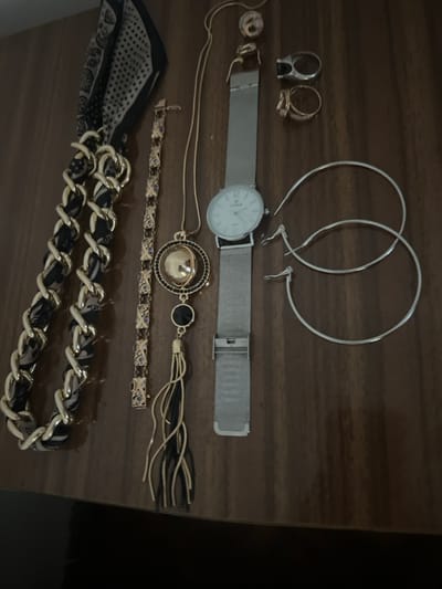 The scene is located in the city of Smederevska Palanka Municipality, Serbia.

An assortment of fashion accessories is displayed on a dark brown, reflective, polished wooden surface. From left to right:
On the far left, a folded fabric item, possibly a scarf or headband, features a black and beige/white geometric or paisley pattern with a distinct black border. Adjacent to it, a large, gold-toned chain accessory, likely a necklace or bracelet, has black fabric interwoven through its links. To its right, a gold-toned link bracelet is visible, accented with small, dark blue or purple-colored stones set within rectangular links.

Further to the right, a long gold-toned necklace is laid out, featuring a complex pendant composed of a spherical gold-toned element encased by a ring of black stones, followed by a smaller black circular disc, and terminating in a tassel made of gold and black strands. Immediately to the right of the necklace, a wristwatch with a silver-toned mesh band and an open clasp is visible. The watch face is white with black numerals or markers and displays the brand "CEL MARE".

Above the watch, two rings are present: one gold-toned ring with a patterned band and another silver-toned ring featuring a black rectangular inlay. To the right of these, a pair of large, silver-toned hoop earrings are laid flat. Below these, two individual small silver-toned earring clasps or hooks are also present.

No individuals are visible in the scene. The objects are stationary, with no actions or interactions depicted.