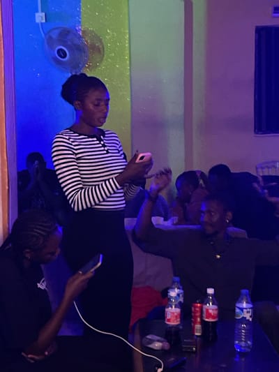 In Durumi, Nigeria, a dimly lit room, possibly a bar or gathering space, is illuminated by blue and green ambient lighting. A woman in a black and white striped long-sleeved shirt and black skirt stands centrally, holding a mobile phone in her right hand and appearing to interact with it. To her left, another woman with braided hair is visible from the shoulders up, also looking down. In the background, several individuals are present, their faces largely obscured by shadow and the low lighting. A small fan is mounted on the blue-lit wall above the central figure. On a table in the foreground, several bottles of water and cans of soda are arranged. The overall atmosphere suggests a casual social gathering.