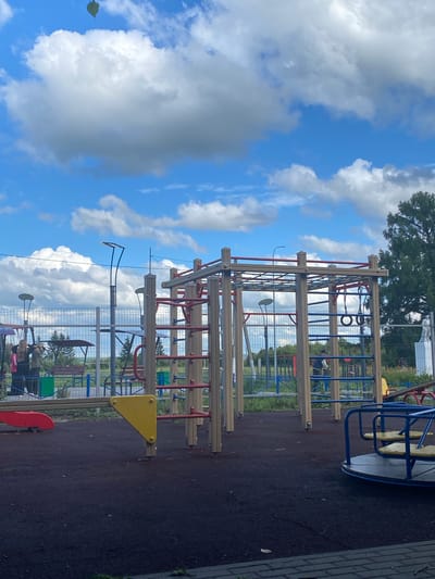This image shows an outdoor playground in Nadezhdino, Russia. The playground features a complex climbing structure with wooden beams and red and blue bars, along with monkey bars and rings. A yellow triangular climbing element is attached to the structure, and a red slide is partially visible. A merry-go-round with yellow seats and blue railings is also present. The ground is covered with dark, soft material. In the background, a fence separates the playground from a grassy area with trees. The sky is bright blue with fluffy white clouds, suggesting it is daytime and the weather is pleasant. A single green leaf is visible floating in the air on the left side of the frame. There are a few people in the background, but they are not clearly discernible. The overall scene depicts a public recreational space on a clear day.