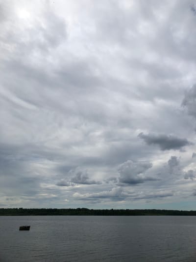 The image depicts a wide shot of a calm body of water, likely a river or lake, under a heavily overcast sky. The water is dark and reflects the muted tones of the clouds. In the distance, a dense, dark green tree line forms the horizon, suggesting a forested area. A small, dark, rectangular object, possibly a floating debris or a small raft, is visible in the water in the left portion of the frame. The sky is filled with various shades of grey clouds, with some lighter patches indicating diffused sunlight. The scene is tranquil and appears to be a natural landscape. The location is the Neva River in Svirstroy, Russia.