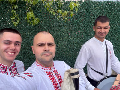 Three men are depicted lying on their backs, with their heads positioned towards the bottom of the frame and their bodies extending upwards. They are set against a backdrop of a dense, dark green artificial hedge or fence designed to mimic foliage. In the lower part of the image, in front of the hedge, natural green leafy branches and vines are visible.
The man on the far left has his eyes closed and a slight smile. The central man looks directly at the camera with a neutral expression. The man on the far right is smiling broadly, revealing his teeth.
All three men are dressed in traditional white long-sleeved shirts, prominently featuring rich red and black embroidered patterns, likely of geometric and floral motifs, along the collar and front placket. These shirts are characteristic of Bulgarian folk attire.
Next to the upper body of the man on the far left, a large, grey, shallow drum (possibly a tupan or davul) with a black strap is partially visible, along with a piece of patterned fabric. Adjacent to the central man's chest is a traditional Bulgarian bagpipe (gaida), identifiable by its dark wooden or horn chanter (gajdunka) and a light-colored fabric bag (meh).
The men appear to be posing for a photograph, likely showcasing traditional Bulgarian folk costumes and musical instruments, suggesting an association with folk music or cultural performance. The overall mood is positive, with smiles ranging from subtle to broad. The bright, even lighting indicates it is daytime. The scene is situated in Rakovski, Bulgaria.