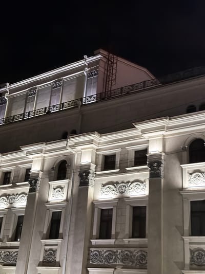 The image shows a low-angle view of a building at night. The building features classical architectural details, including decorative moldings, window frames, and balconies. The building's facade is lit, highlighting its ornate design. The background is a dark night sky. The location cannot be determined from the image.