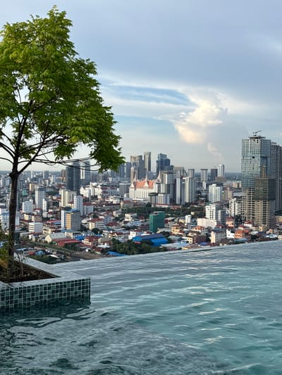 A rooftop infinity pool overlooks a dense urban landscape in Khan Mean Chey, Cambodia. The pool's edge is tiled with small, dark green and white square tiles, and the water reflects the sky and surrounding buildings. In the foreground, a large tree with vibrant green foliage partially obscures the view. The cityscape is a mix of residential buildings with red and blue roofs, as well as modern high-rise structures, some of which appear to be under construction. The sky above is partly cloudy, with a large white cloud formation dominating the upper portion of the image. The lighting suggests it is either early morning or late afternoon. No people are visible in the image.