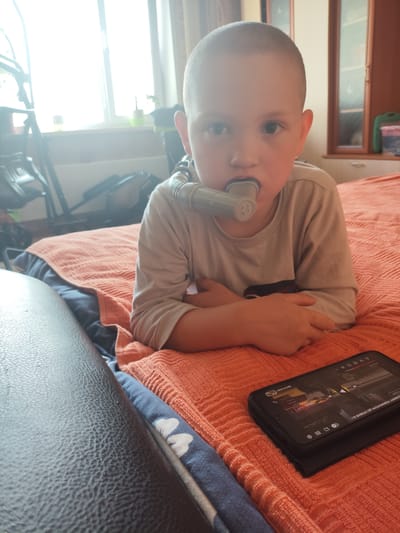 A young boy with a shaved head is lying on an orange blanket, wearing a light-colored t-shirt. He has a medical device with a tube entering his mouth. His arms are crossed in front of him, and he is looking at the camera. In the foreground, a smartphone is lying on the blanket, displaying a video with Ukrainian text. The background shows a window with bright daylight, a piece of furniture with glass doors, and a stroller. The scene appears to be indoors, possibly in a bedroom, and no specific location in Askiz, Russia, can be determined from the image.