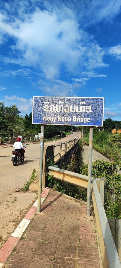This is a medium shot of a bridge in Xayden, Laos. The bridge is made of concrete with a metal railing. There is a blue sign with white lettering that says "Houy Keua Bridge" in English and Lao script. A person on a motorcycle is riding across the bridge. The sky is blue with some white clouds. The bridge is surrounded by lush green vegetation.
