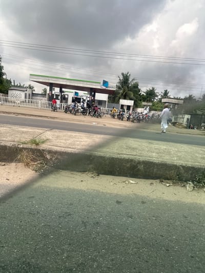 The image captures a roadside scene at an NNPC branded gas station in Igwuruta, Nigeria, under an overcast sky. The gas station features a white canopy with green and white trim, covering multiple fuel pumps. A white metal fence separates the gas station forecourt from the adjacent dirt/gravel shoulder and asphalt road.
Approximately 15-20 motorcycles are queued or parked on the dirt forecourt in front of the gas station, primarily positioned to the left and center. Several individuals, around 10-15, are standing near the motorcycles and fuel pumps. One person in a red shirt stands next to a motorcycle on the far left. In the mid-ground, on the right side of the image, a male figure dressed in a light-colored traditional garment walks from right to left across the dirt shoulder, moving away from the gas station.
The immediate foreground shows an asphalt road surface with a raised concrete curb running parallel to it. Sparse dry vegetation and scattered debris are visible along the curb and dirt shoulder. Power lines traverse the upper portion of the frame. The background includes various green trees, including palm trees, and additional indistinct white structures.