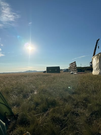 The image depicts a wide, grassy field under a bright, sunny sky with some wispy clouds. In the foreground, a green tent is partially visible. In the middle ground, a dark green, shed-like structure with windows stands to the left. Further to the right, a flatbed truck is parked, carrying a large wooden lattice structure. A crane is attached to the truck, and a large, white tarp-covered object is situated next to it. In the distance, rolling hills and wind turbines are visible. The sun is high in the sky, creating a lens flare effect. The scene suggests an outdoor work or event setup in a rural, possibly remote, location.