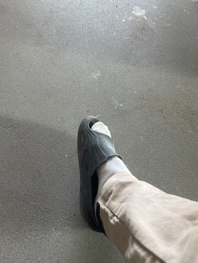 The image shows a close-up, overhead view of a person's left leg, clad in a light-colored, loose-fitting pant and a white sock. The leg is encased in a dark gray slide sandal. The scene appears to be outdoors, on a textured, dark gray surface, possibly asphalt or concrete. The lighting suggests it is daytime. The location is Strumivka, Ukraine.