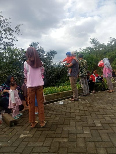 This outdoor photograph captures a group of people, including several adults and children, gathered on a paved area with green foliage and distant hills in the background, likely in Pangalengan, Indonesia. The sky is overcast but bright, indicating daytime.
In the foreground and center, a young person, possibly a teenager or young adult, stands with their back to the camera, wearing a brown hijab, a pink long-sleeved top, and brown cargo pants. To their left, a woman sits on a low stone ledge next to a small child in a pink dress and white top, who is looking down. Another child is partially visible behind them.
Further to the right, a man with a light-colored cap holds a baby wearing a red top and blue hat, both also facing away from the camera, looking towards the green landscape. Behind them, another man stands. Several other individuals are seated further to the right along a raised plant bed. One woman, wearing a grey hijab, appears to be holding a red object, possibly a balloon or ball. Another person sitting wears a red shirt.
The ground is covered with grey interlocking paving bricks. Lush green trees and bushes form the mid-ground and background, with hints of hills or mountains visible in the distance through the vegetation. The overall atmosphere appears casual and relaxed, suggesting a family outing or leisurely gathering in a scenic outdoor location. There is no visible text in the image.