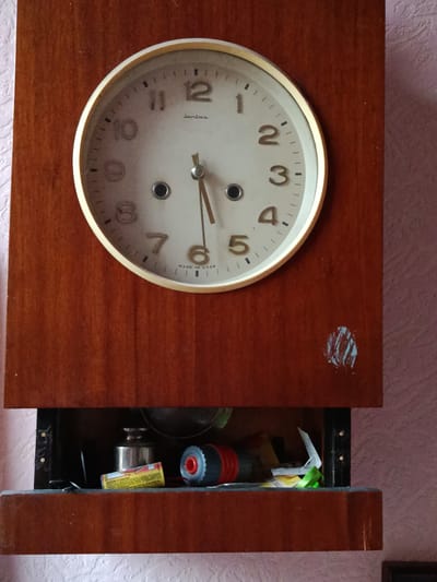 A wooden mantel clock with a white face and gold-colored trim is mounted on a textured pink wall. The clock features black Arabic numerals for hours and black tick marks for minutes. The hands are thin and metallic, with the hour hand pointing approximately to the 11 and the minute hand pointing to the 5. The brand name "Janus" is visible above the center, and "MADE IN USSR" is printed below the 6. Below the clock, there is an open compartment containing various items, including a cylindrical metal object, a yellow and red tube, a black and red cylindrical object, and some paper scraps. The compartment appears to be a drawer or shelf. The overall scene suggests a domestic setting, possibly a home in Смілавіцкі сельскі Савет, Belarus.