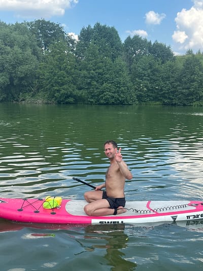 A man is paddleboarding on a calm lake in Dal'nie Prudishchi, Russia. He is seated on a pink and white paddleboard, holding a paddle. He is wearing black shorts and is shirtless. He is smiling and giving a peace sign. The lake is surrounded by lush green trees. The sky is blue with a few white clouds.