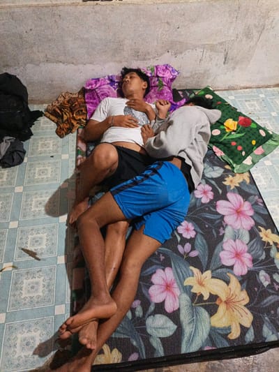 Two individuals are lying on mattresses on the floor in what appears to be a room in Meunasahuket, Indonesia. The room has a concrete wall and a tiled floor. The mattresses are covered with patterned fabrics. The individual on top, wearing a white t-shirt and black shorts, is lying on their back with their eyes closed and one arm resting on their chest. The individual below, wearing a grey hoodie and blue shorts, is lying on their side, facing the other person and with their head obscured. Their legs are intertwined with the person above. Several pieces of clothing and bags are visible on the floor around the mattresses. A patterned pillow with flowers is placed near the heads of the individuals.