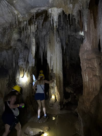 The image depicts two individuals exploring a cave in Khao Phang, Thailand. Both are wearing yellow hard hats, suggesting they are on a guided tour or engaging in spelunking. The cave environment is characterized by rocky formations, stalactites, and stalagmites, with a damp, earthy appearance. Artificial lighting, likely from headlamps or flashlights, illuminates parts of the cave, casting shadows and highlighting the textures of the rock.
The individuals appear to be navigating the cave floor, possibly crawling or moving in a low-slung posture. One person is seen with their legs extended, while the other is in a more crouched position. The overall atmosphere suggests an adventurous activity in an underground setting. The time of day and weather are not discernible from the image, as the scene is entirely within the cave. There are no visible text elements in the image.