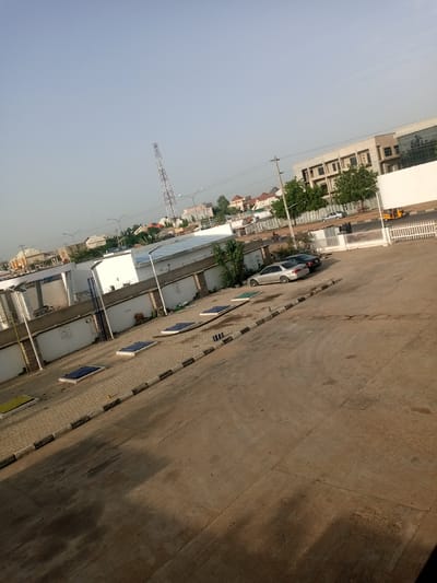 The image captures an outdoor scene with a clear sky and distant buildings. In the foreground, a paved area with distinct square tiles and a concrete surface occupies the majority of the frame. Several rectangular structures with blue covers are embedded within the tiled area. A curb with black and white markings separates the tiled section from the concrete.
In the midground, there is a white-walled building complex with a metal fence. Several cars are parked in front of this complex. Further back, a telecommunications tower is visible against the sky, along with various other buildings, some of which appear residential with pitched roofs. A yellow auto-rickshaw is seen near a white fence on the right side of the image. Green trees are scattered throughout the scene, particularly in front of some of the buildings. The overall impression is an urban environment with a mix of commercial and residential structures. The scene appears to be located in Hotoro North, Nigeria, as indicated by the provided context. There are no people visibly present or engaged in any specific actions. The lighting suggests it is daytime, likely morning or late afternoon due to the angle of the shadows.