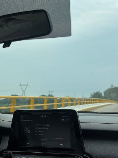 The view from inside a car on a rainy day, presumably in transit, is captured with a focus on the dashboard and the road ahead. The dashboard features a car infotainment system displaying music information and connectivity options like Bluetooth. Through the rain-streaked windshield, a road marking with yellow and black stripes is visible, alongside power lines. The rearview mirror is also in view. The location details, specifically Cucuyulapa, Mexico, are not determinable based on the given image alone.