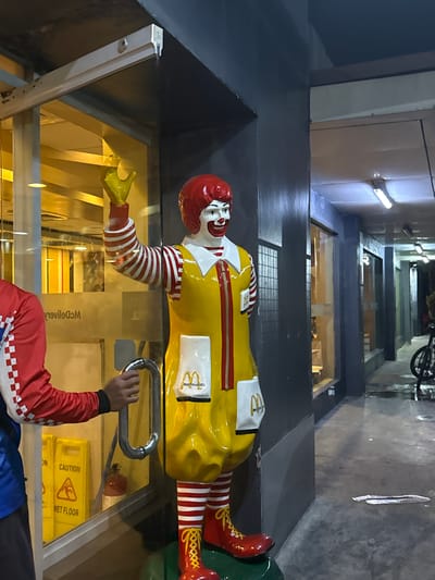 The image shows a statue of Ronald McDonald in front of a McDonald's restaurant at night. The statue is dressed in its iconic yellow jumpsuit with red and white striped sleeves and red shoes. One of Ronald's hands is extended, and the other is holding a silver handle. A person wearing a red jacket with a white checkered pattern and blue pants is visible through the glass, reaching for the same handle. In the background, there are caution wet floor signs and a fire extinguisher. The scene appears to be an outdoor walkway or entrance to the restaurant, with a dark, urban setting. The time of day is night, as indicated by the artificial lighting. The location context is Manila, Philippines.