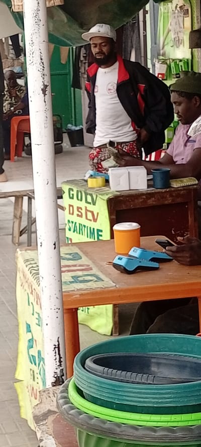 The scene depicts an outdoor commercial or service point in Kwachiri, Nigeria. In the foreground, a white, weathered pole stands, partially obscuring a stack of plastic basins or bowls in various shades of green, black, and gray. To the right of the pole, an orange plastic table supports a vibrant yellow and green banner. The banner displays "GOLV," "DStv," and "STARTIME" in green and red lettering, indicating services offered.
On the table, two blue point-of-sale (POS) terminals are visible, along with an orange cup with a white lid, a yellow roll of tape, and two small white boxes. A man wearing a pink shirt and a dark green cap is seated behind this table, holding a mobile phone and looking down at it.
Standing to the left of the seated man is another man, dressed in a white t-shirt, a black jacket with red trim, white baseball cap, and red-patterned shorts. He holds a stack of cash in his hands and is looking downwards. In the deeper background, another individual wearing a patterned shirt is seated on an orange plastic chair, partially visible. The background also reveals a green awning or tarp overhead, a green wall or doorway, and what appears to be merchandise or clothing hanging. A black bucket is visible on the ground.