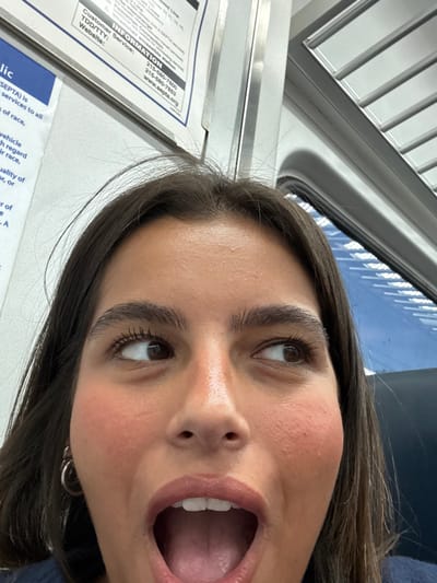 The image captures a close-up selfie of a young adult female inside a SEPTA public transit vehicle, specifically a train or subway car, in Philadelphia, United States. The female subject has dark brown hair and brown eyes, looking upwards and to her right. Her mouth is wide open, exposing her teeth and tongue. A silver hoop earring is visible on her left ear. The background on the left features a white information panel with "SEPTA Customer Service" text, phone numbers (215-580-7800), and "www.septa.org". On the right, a window reveals a blurred blue exterior view with horizontal reflections, indicating motion. The vehicle's light gray interior ceiling structure is partially visible above the window.