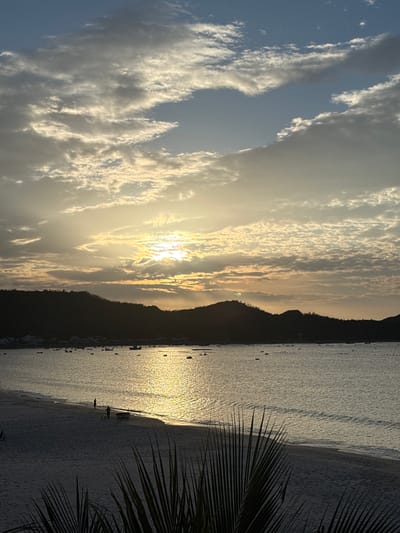 A vertical, eye-level shot captures a serene sunset over a coastal scene in Hòa Hiệp, Vietnam. The sun, partially obscured by wispy clouds, casts a golden glow on the water, creating a shimmering reflection that stretches towards the shore. Silhouetted against the vibrant sky are rolling hills covered in vegetation, with a small town nestled at their base. Numerous small boats are scattered across the calm sea. The foreground is dominated by a sandy beach, with two individuals walking near the water's edge and a small boat pulled ashore. Palm fronds in the lower right corner frame the scene. The sky is a blend of soft blues and grays, with dramatic cloud formations illuminated by the setting sun.