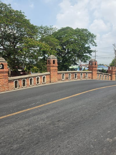 A brick wall and gate run alongside a road in the Historic City of Ayutthaya, Thailand. The wall features arched openings and turrets at intervals. The road has a yellow center line. Trees line the other side of the wall. Overhead power lines are visible. The sky is lightly cloudy.