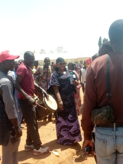 The image depicts an outdoor gathering under bright sunlight in Kagoma, Nigeria. The ground is reddish-brown dirt. In the foreground, a man wearing a red shirt, dark pants, and white sneakers plays a djembe-style drum with a stick. To his right, a woman in a long, dark purple patterned dress, dark headwrap, and large sunglasses smiles, holding a clear plastic water bottle in her left hand and a dark smartphone in her right. Gold-colored bracelets are visible on her left wrist. To the left of the drummer, a man in a red baseball cap and light-colored top is partially visible. The back of another man, wearing a brown long-sleeved shirt, blue jeans, and a camouflage shoulder bag, occupies the right foreground, holding a dark smartphone and keys in his right hand. A crowd of individuals in various attire is visible in the mid-ground and background. A light-colored building with a white roof and some trees are discernible in the distant background. A small, white, elongated object rests on the ground near the drummer's feet.