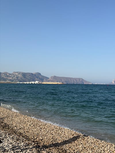 The image depicts a coastal view in Altea, Spain, on a clear, sunny day. In the foreground, a pebble beach stretches across the bottom left of the frame, with gentle waves lapping at its edge. The sea, a deep teal color, occupies the middle ground, showing moderate choppiness. In the background, a sprawling cityscape is nestled against a mountainous backdrop. Buildings with white and light-colored facades are visible, indicating a densely populated area. Several boats are docked in a harbor to the left of the city. The distant mountains are rugged and brown, with some areas appearing to be terraced or built upon. The sky is a clear, pale blue, devoid of clouds. The lighting suggests it is daytime, likely mid-morning or afternoon, given the bright sunlight and clear visibility. There are no people or animals visible in the frame. The scene is static and serene.