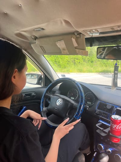 The image depicts the interior of a Volkswagen car, viewed from the passenger's perspective. A young woman, with dark hair and a black shirt, is seated in the driver's seat. Her hands are on the steering wheel, which is adorned with a blue and black patterned cover. The dashboard displays the car's instrumentation, including the speedometer and tachometer. To the right of the steering wheel, in the center console, a can of Coca-Cola is placed in a cup holder. The exterior environment visible through the windows is a sunny day with green foliage, suggesting a rural or suburban setting. The interior of the car shows some wear, with a sagging headliner visible towards the top of the frame. There is an air freshener hanging from the rearview mirror. The car appears to be parked. The location is Vlahovo, Bulgaria.