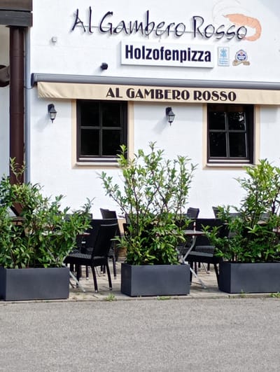 The image shows the exterior of a restaurant named "Al Gambero Rosso" in Brannenburg, Germany. The restaurant's facade is white with dark trim around the windows. A beige awning extends over the windows, with the restaurant name "AL GAMBERO ROSSO" printed in large dark letters. Above the awning, the restaurant's name "Al Gambero Rosso" is also displayed in stylized lettering, accompanied by a graphic of a shrimp. Below this, a sign reads "Holzofenpizza". Two small framed emblems are visible to the right of the "Holzofenpizza" sign. In front of the restaurant, there are outdoor seating arrangements, with tables and chairs, partially obscured by large planters filled with green shrubs. The scene appears to be captured during daylight hours.