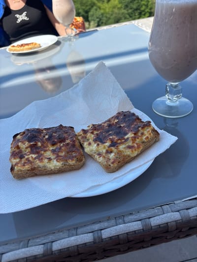The scene is an outdoor setting, possibly a patio or balcony, in daytime. A grey surface, likely a table, holds a partially filled glass with a light pink smoothie and a white plate containing two pieces of toast with a melted topping that appears browned and slightly burnt. The toast rests on a white paper napkin. To the left, a reflection on the table shows another person sitting at the table with a black shirt with a small white flower, and a plate with a sandwich with melted topping on it. A woven grey structure, perhaps part of outdoor furniture, is seen along the right edge of the image. The green vegetation and blue sky are visible through reflection in the table. The location is Tserovo, Bulgaria.