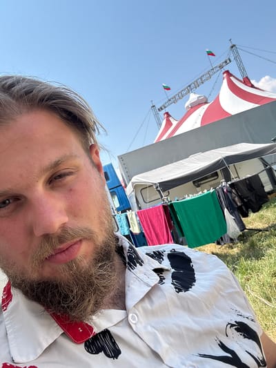 A selfie taken outdoors in Kavarna, Bulgaria, features a man with blond hair and a beard in the foreground. He is wearing a white shirt with a black and red abstract pattern. In the background, a red and white striped circus tent is visible under a clear blue sky. Two Bulgarian flags are flying from the tent's structure. A caravan and a clothesline with laundry are also present, suggesting a temporary living situation, possibly related to the circus. The ground is covered in grass.