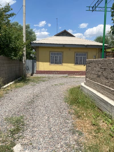 The image depicts a single-story yellow house with a corrugated metal roof, situated in a rural or suburban area. The house has two visible windows with white frames and translucent curtains. A decorative red base runs along the lower part of the house's exterior wall. The area in front of the house is a gravel driveway. To the left of the driveway is a concrete block wall, and further to the left are trees. A utility pole is visible behind the concrete wall. To the right of the driveway is a low, weathered brick structure with a concrete base, and some dry and green grass. A green metal support structure, possibly for a clothesline or awning, is visible against a bright blue sky with scattered white clouds. A thin metal fence or gate is seen between the concrete wall and the house. The overall scene suggests a sunny day. The location of Боғистон, Tajikistan is indicated, suggesting this is a property within that region. There are no people or animals visible in the image.