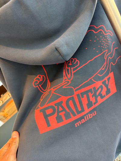 The image shows a close-up of a dark gray hooded sweatshirt with a bright red graphic. The graphic features stylized lettering that spells out "PASTRY" in large, bold, uppercase letters, with "malibu" in smaller, lowercase letters beneath it. The lettering is angled, suggesting movement. To the left of the text, there is a red outline of what appears to be a hand or paw with a fist raised, and to the right, there is a wavy line and a cluster of red dots, possibly representing an explosion or a design element. A part of the sweatshirt's hood is visible on the left side of the frame. The fabric of the sweatshirt is textured with visible knitting. The background is out of focus but suggests an indoor setting with some shelves or displays visible. The photograph is taken from a slightly elevated angle, focusing on the graphic on the back of the sweatshirt. There is no indication of the specific location within Malibu, California.