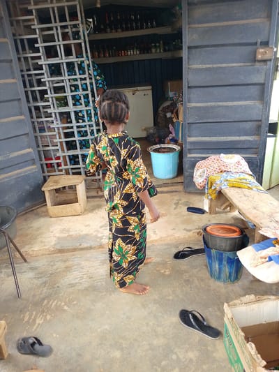 The image depicts a young girl standing in front of a shop in Iseyin, Nigeria. She is facing away from the camera, wearing a colorful long dress with a black base adorned with green and yellow leaf-like patterns. Her hair is styled in braids. To her left is a metal shelving unit, and to her right, a dark-colored shuttered door is partially open, revealing shelves stocked with bottles and other items inside the shop. 

In front of the shop, there is a light-colored concrete floor. Various objects are scattered around. To the girl's left, there is a small wooden stool with an open compartment. A large grey basin is visible to the far left of the frame, with a metal stand next to it. To the girl's right, a blue plastic bucket sits on the ground, filled with dark liquid. On top of it is another bucket with an orange lid. Further to the right, a wooden bench is covered with brightly patterned fabrics, and a pair of dark blue flip-flops lies on the ground in front of it. Another pair of black flip-flops is on the floor in the lower right of the image, next to an open cardboard box. 

The scene appears to be outdoors, likely during the daytime, with neutral lighting. The overall impression is one of a busy market or shop environment.