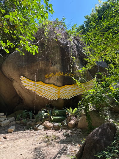 The image depicts a large rock face in Hoa Thang, Vietnam, adorned with a painted yellow and white wing motif and text in yellow lettering that reads "ĐẢO HOA LAN". The rock is situated in a natural environment with lush green foliage and trees overhead, suggesting an outdoor location, possibly a tourist attraction or a natural landmark. The ground in front of the rock is covered with dirt, pebbles, and some scattered stones. In the lower portion of the image, there are several green steps leading up to a moss-covered area in front of the rock, possibly part of a pathway or an altar-like structure. The lighting indicates a sunny day with shadows cast by the surrounding vegetation. No people are visible in the frame.