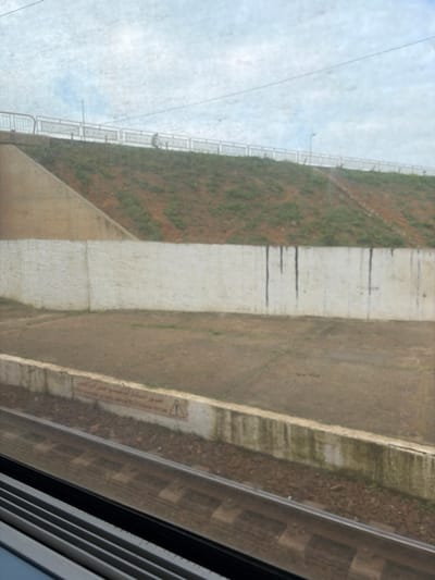 A sideways view from a train window shows a landscape passing by. A grassy embankment is to the left, reinforced with a concrete wall and a guardrail at the top. A wider concrete area, likely a pathway, runs parallel. Train tracks are visible closer to the window, alongside a warning sign written in Arabic. The sky is cloudy. The image has a slightly faded or blurred quality.
