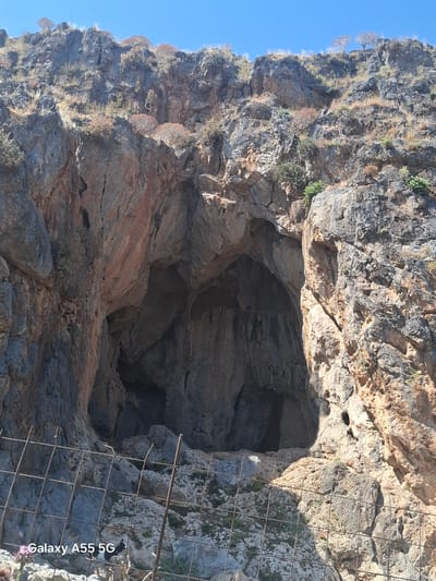 The image captures the entrance of a large cave set into a cliff face in Plakias, Greece. The cliff is composed of rugged rock formations, with varying colors from light tan to dark gray. Some sparse vegetation clings to the rock face. A wire fence is partially visible in the foreground, potentially restricting access to the cave. The sky is a clear blue. The entrance of the cave is dark, suggesting a significant depth to the cavern.