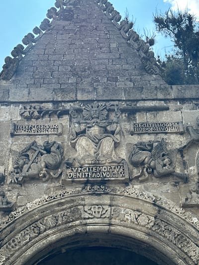 The image shows a detailed stone carving above an arched entrance, likely part of the San Andrés Apóstol church in San Andrés Calpan, Mexico. The carving depicts a central seated figure, possibly Christ or a deity, with a radiant halo. Flanking this central figure are winged beings and inscriptions in Latin. The left side shows a winged figure holding a cross, and the right side shows another winged figure. The stonework is weathered and shows signs of age, characteristic of historical architecture. The surrounding environment is partially visible, with the sky and some foliage suggesting an outdoor setting. The overall scene is one of religious iconography carved into stone.