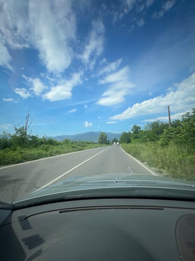 The image is taken from the interior of a car, looking out through the windshield. The car is traveling on a two-lane asphalt road that curves to the right. The road is bordered by lush green vegetation and trees on both sides. In the distance, a range of mountains is visible under a bright blue sky dotted with white clouds. The hood of the car, a light gray Mercedes-Benz, is visible in the foreground. The car appears to be in motion, as evidenced by the slightly blurred roadside vegetation and the dynamic perspective. There are several white vehicles visible in the distance on the road. A wooden utility pole is present on the right side of the road. The scene is set in a rural or mountainous area, consistent with locations around Mezdreya, Bulgaria.