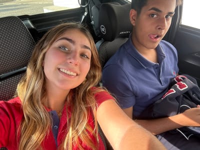 The image depicts two individuals seated inside a vehicle, located in Lordelo do Ouro e Massarelos, Portugal. In the left foreground, a female subject with long wavy blonde hair, heterochromatic blue-green eyes, and a fair complexion smiles directly at the camera. She wears a red polo shirt with a navy blue collar and placket, along with a gold-colored pendant necklace. To her right and slightly behind, a male subject with dark hair and a darker complexion is seated. He is looking towards the camera with his tongue slightly extended in a playful manner. He wears a navy blue polo shirt with a small embroidered emblem and holds a dark blue jacket or sweatshirt featuring white and red lettering. The vehicle's interior shows dark grey patterned fabric on the seats, and a headrest with a "NIRACP+" logo is visible behind the male subject. Bright natural light enters the vehicle from the upper right.