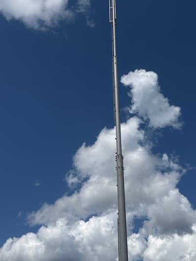 A tall, silver antenna tower stands against a bright blue sky dotted with white, fluffy clouds. The tower is segmented and appears to be constructed of metal, with visible bolts and joints along its length. The bottom of the tower is obscured by a large cloud formation, while the top extends upwards, displaying a series of vertical antennas. The image is taken from a low angle, looking up at the tower and sky. There are no people or animals visible in the scene. The location is identified as Schokino, Russia.