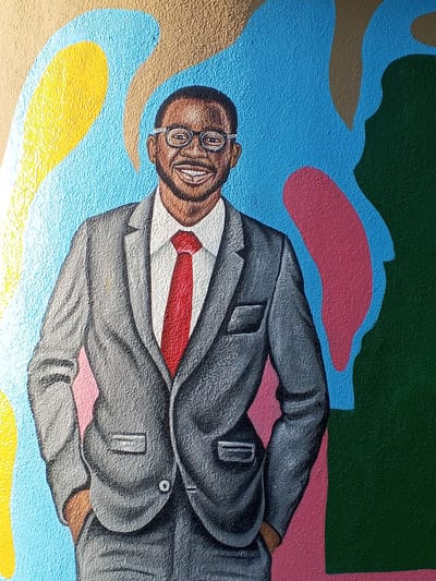 A mural in Tawpo, Nigeria, depicts a Black man wearing a grey suit, white shirt, and red tie. He has glasses, a beard, and is smiling. His hands are in his pockets. The background consists of various shapes and colors, including yellow, blue, pink, and green. The wall has a textured surface.