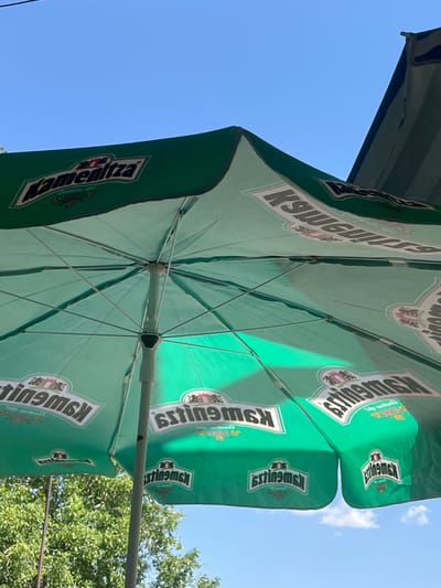 The image shows the underside of two green outdoor umbrellas with branding. The umbrellas are open and angled towards the sky, which is blue with some clouds. Sunlight filters through the fabric, casting shadows and highlighting the umbrella's structure of metal poles and ribs. The branding on the umbrellas appears to be in white text with a pink outline, and includes what looks like a crest or logo with intricate details. Visible text on the umbrellas includes "Kamenitza" on one and repeated instances of "Brlinsmsk" or similar lettering on both. The setting appears to be outdoors, possibly at a cafe or outdoor seating area, with green foliage from trees visible in the upper right corner. The overall impression is a bright, sunny day.